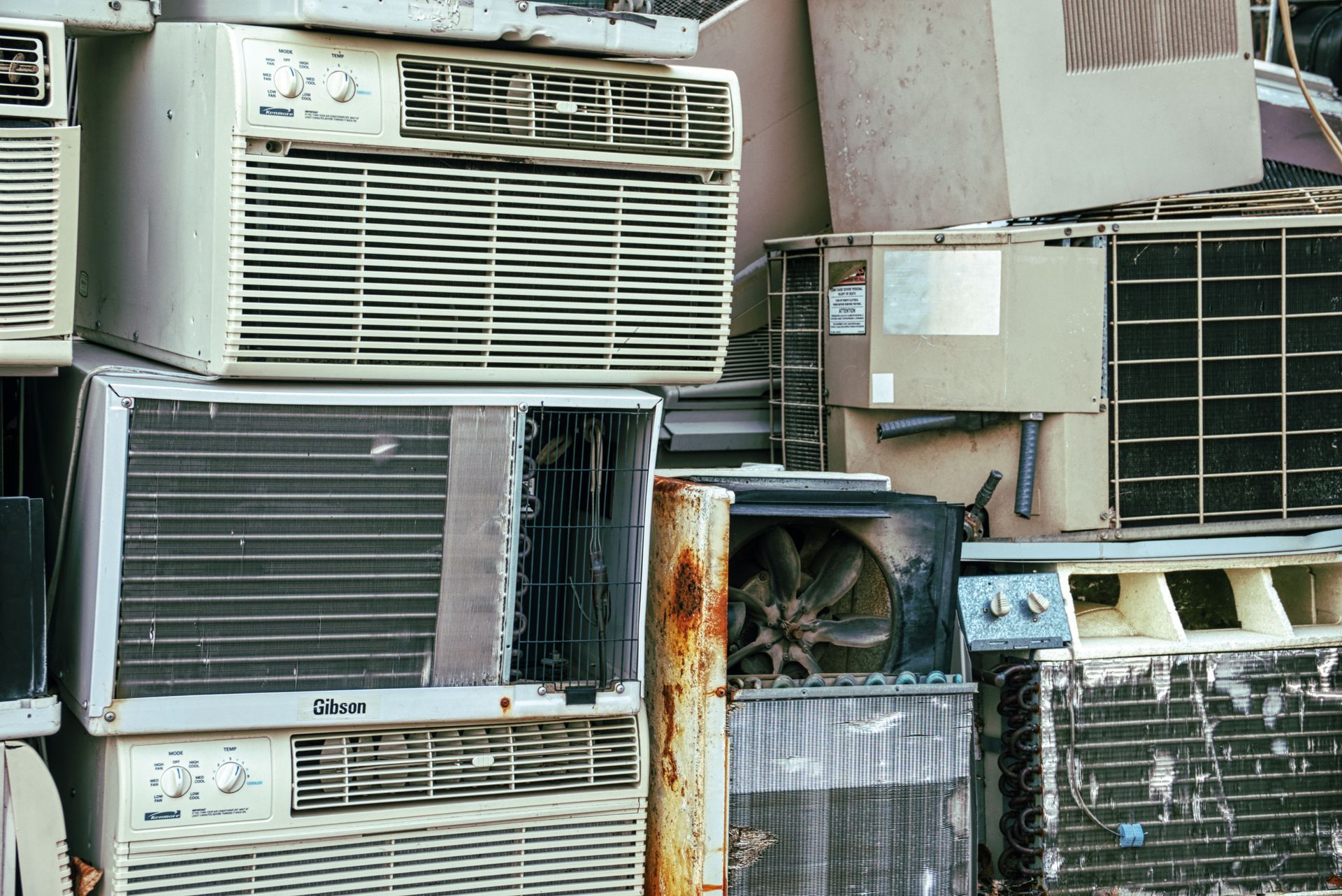 A collection of broken and rusted air conditioners piled in an outdoor junkyard.